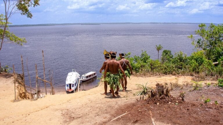 Indigenous men in traditional attire overlooking boats on the Amazon River.