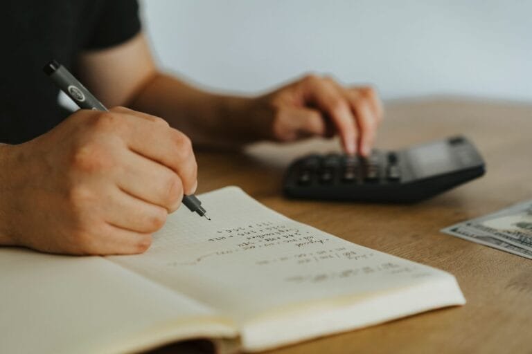 Close-up of hands writing calculations in a notebook with a calculator, focused on budgeti