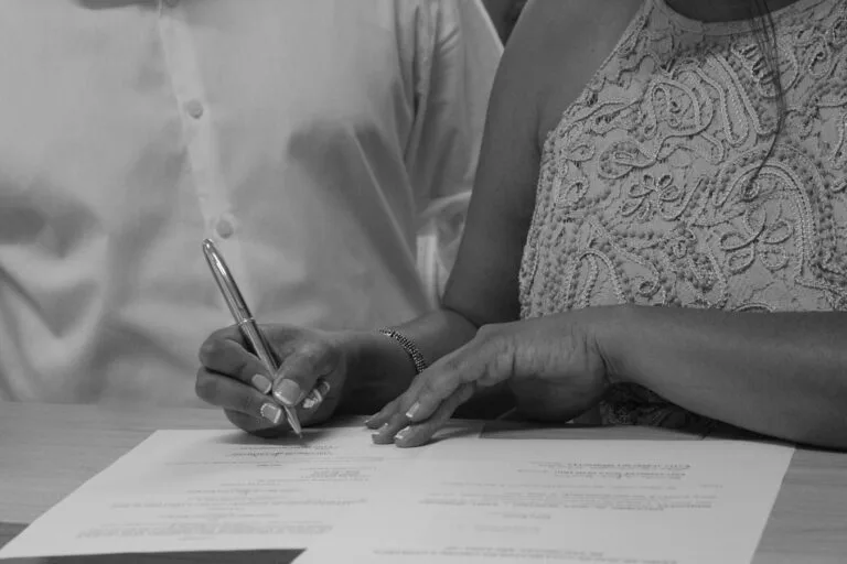 Black and white image of a couple signing documents, focusing on their hands and pen.