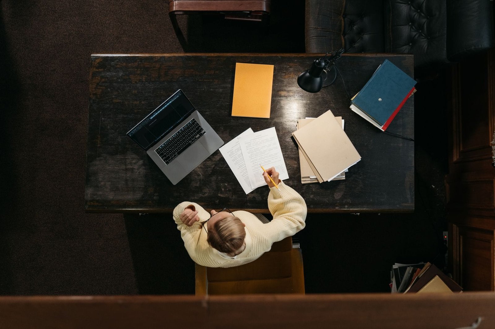 Person in yellow sweater working at a wooden desk with documents, folders, and a laptop.
