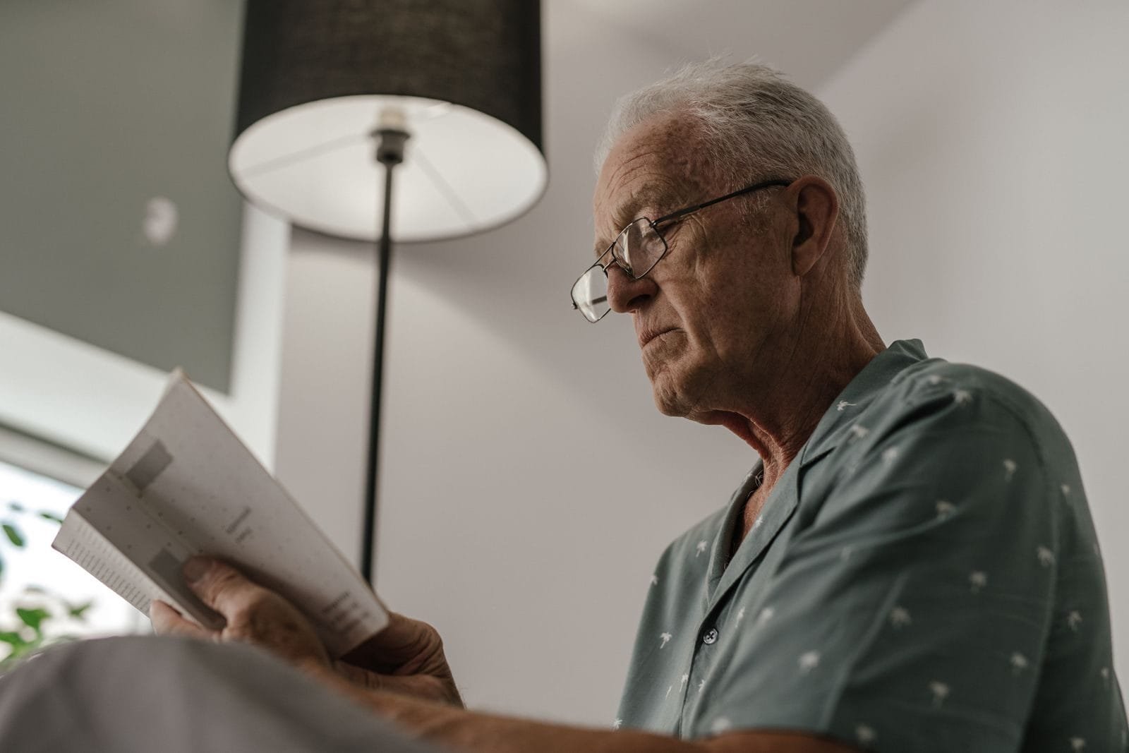 Reajuste do INSS: Índice Usado e Quando o Valor Muda 1 Senior man with eyeglasses reading a book under a lamp in a comfortable and serene indoor setting.