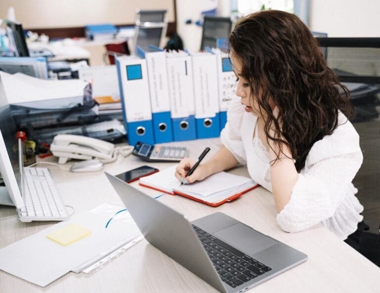 Como podemos ajudar? 14 An office worker takes notes while working at a desk with a laptop and documents.
