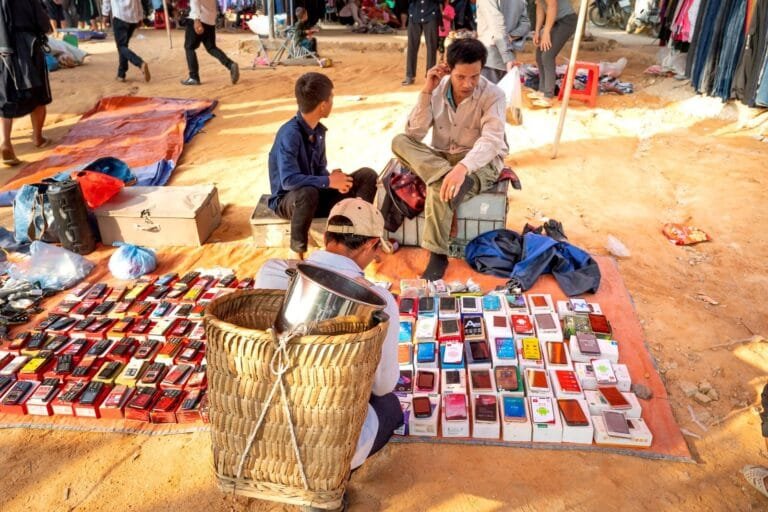 From above of anonymous ethnic male locals sitting on chairs on sandy ground and selling various smartphones on bazaar