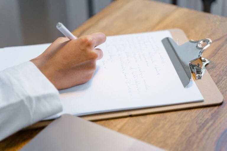 A close-up of a persons hand writing on a clipboard on a wooden table, displaying focus and concentration.