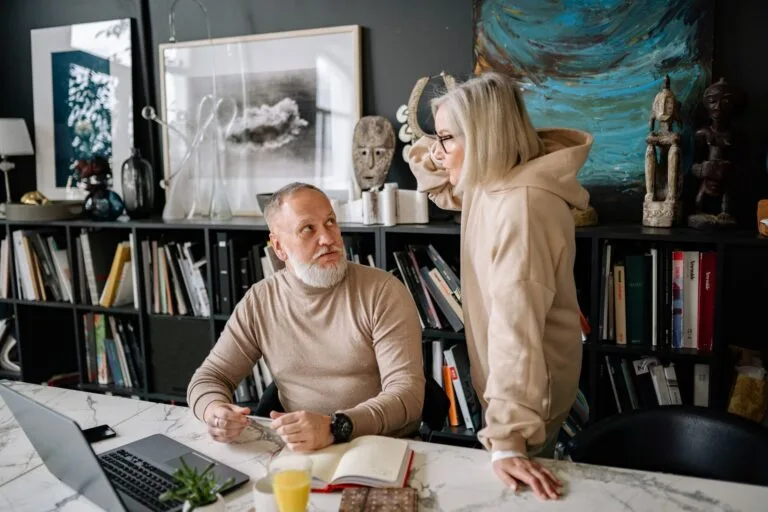 Elderly couple discussing something at a marble table with books and laptop, surrounded by