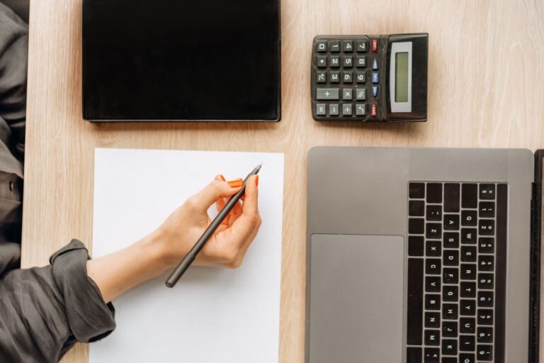 A person writing on paper surrounded by a laptop, tablet, and calculator on an office desk