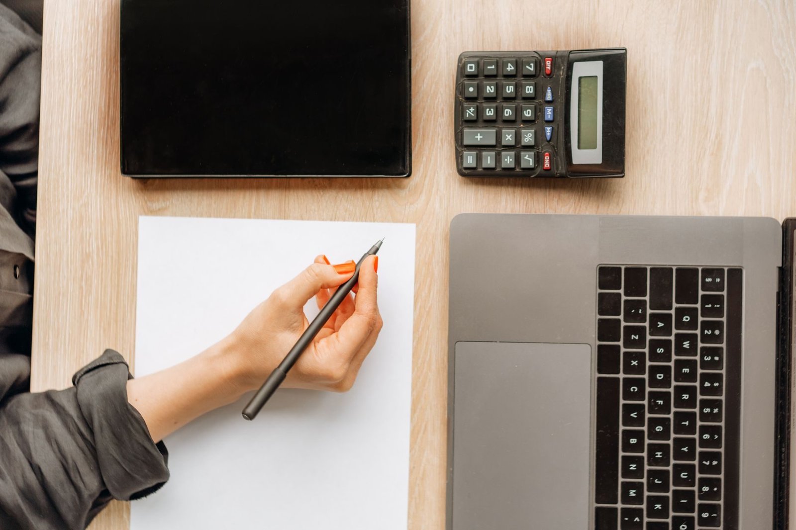 A person writing on paper surrounded by a laptop, tablet, and calculator on an office desk