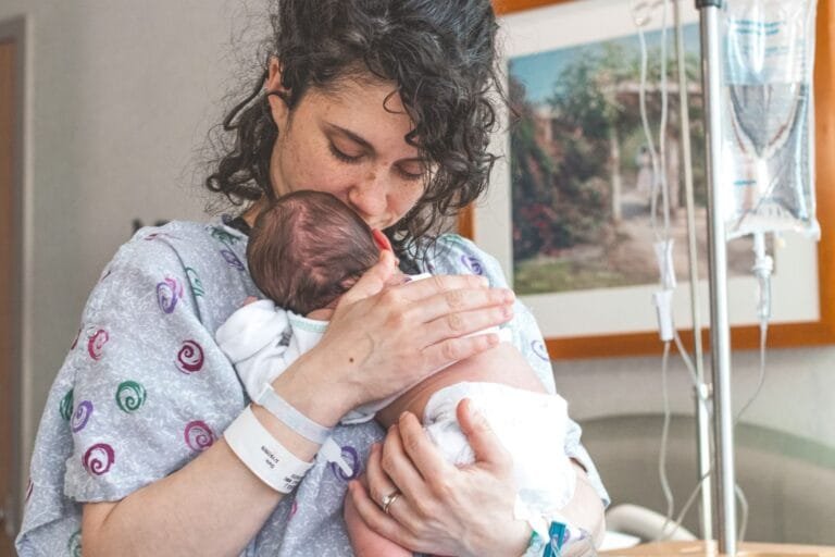 Salário-Maternidade 2026: Quem Tem Direito, Duração e Valor 4 Mother gently holding newborn baby in hospital setting, showcasing warmth and love.