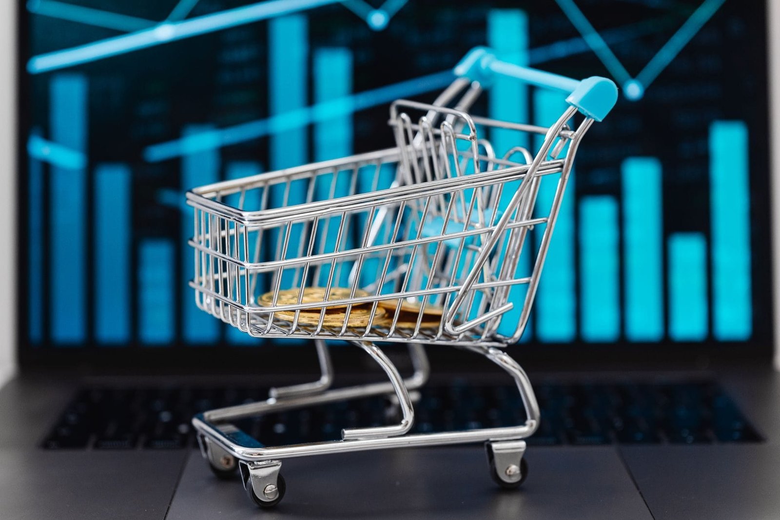 A miniature shopping cart with coins sits on a laptop displaying financial graphs, symboli