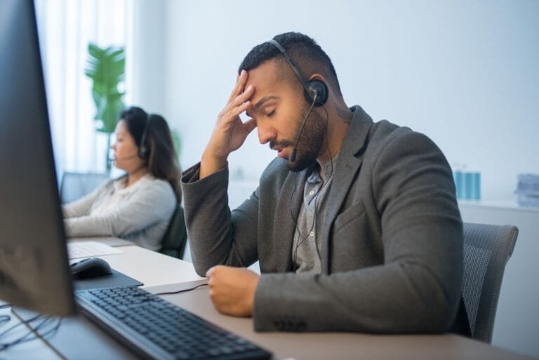 A tired call center employee with a headset looks stressed at his desk in a modern office.