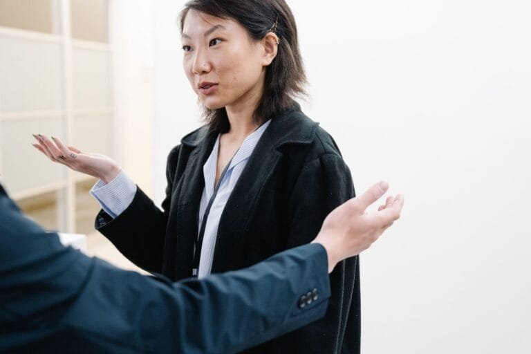Asian woman in conversation with gestures, wearing a black blazer, indoor setting.