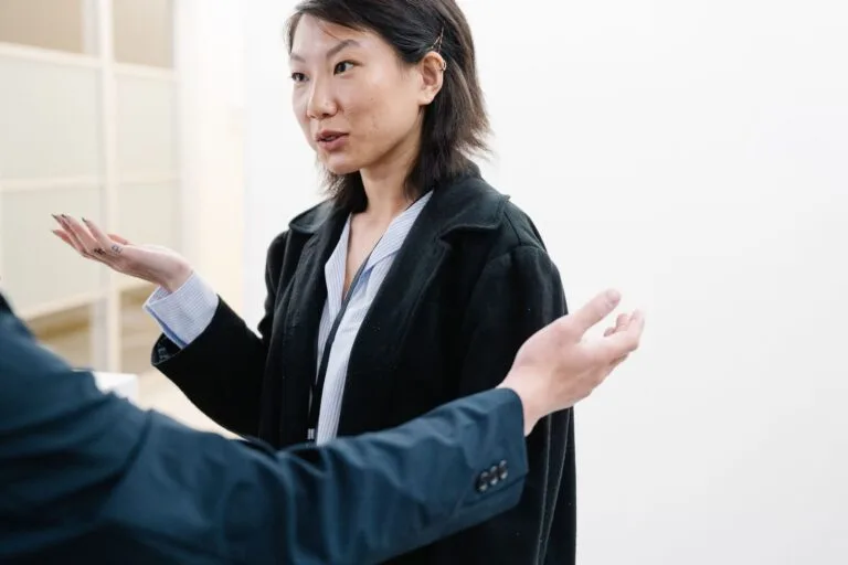 Asian woman in conversation with gestures, wearing a black blazer, indoor setting.
