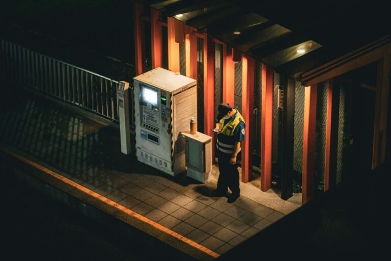 A security guard checks his smartphone beside an illuminated vending machine at night.