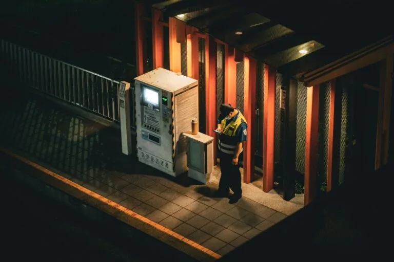 A security guard checks his smartphone beside an illuminated vending machine at night.