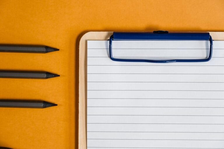 Top view of a lined clipboard with pencils on a vibrant yellow background.