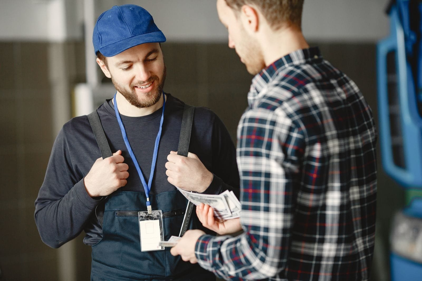 Tabela de contribuição do INSS de 2026 mantém alíquotas progressivas de 7,5% a 14% em quatro faixas salariais 1 A mechanic in a blue cap receives cash payment from a customer in a plaid shirt, smiling.