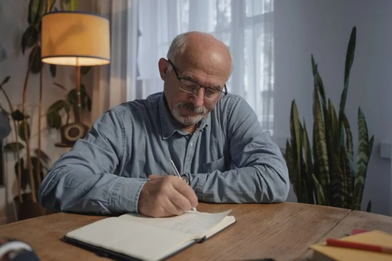 A senior adult man writing in a notebook at a desk in a cozy home office setting.