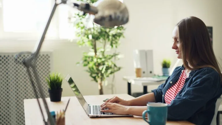 Woman smiling while working on a laptop in a modern office with plants.