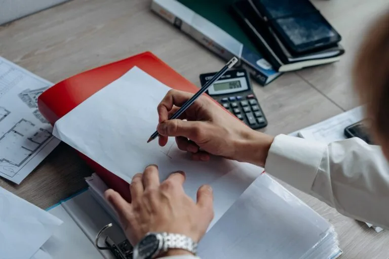Close-up of hands writing on paper at a desk with a calculator, notebook, and drawing plan