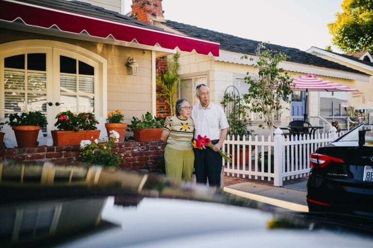 An elderly couple stands in front of a charming house holding a bouquet, capturing a serene lifestyle moment.