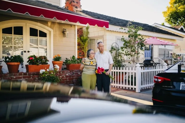 An elderly couple stands in front of a charming house holding a bouquet, capturing a serene lifestyle moment.