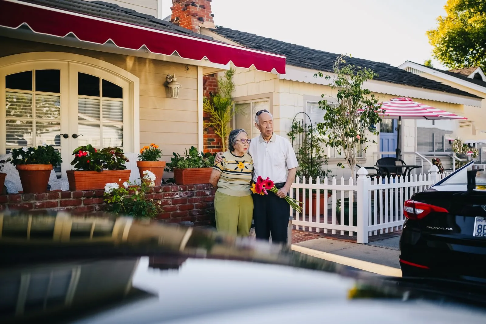 An elderly couple stands in front of a charming house holding a bouquet, capturing a serene lifestyle moment.