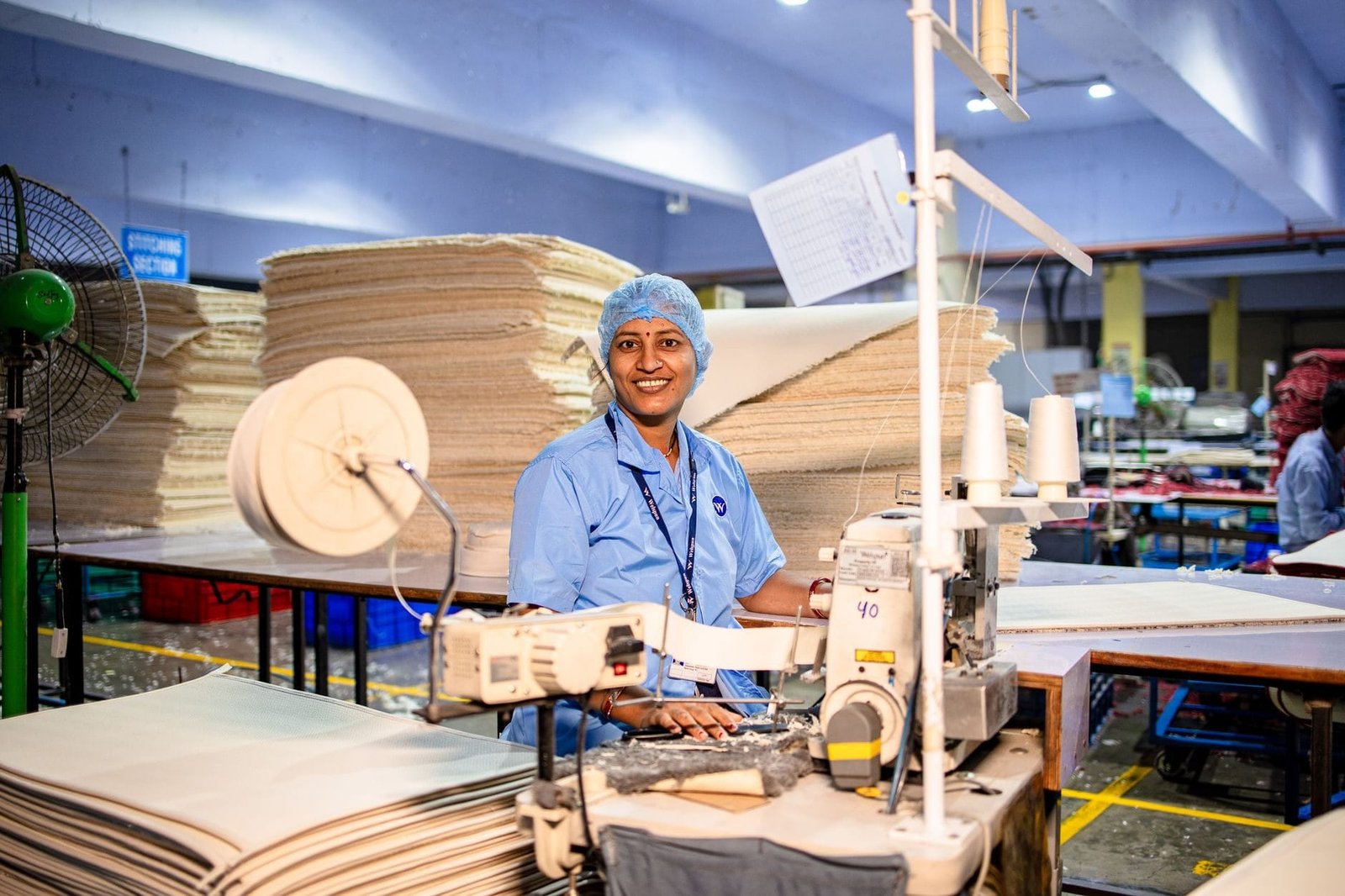 Happy female textile worker in factory, operating a sewing machine with stacks of fabric.