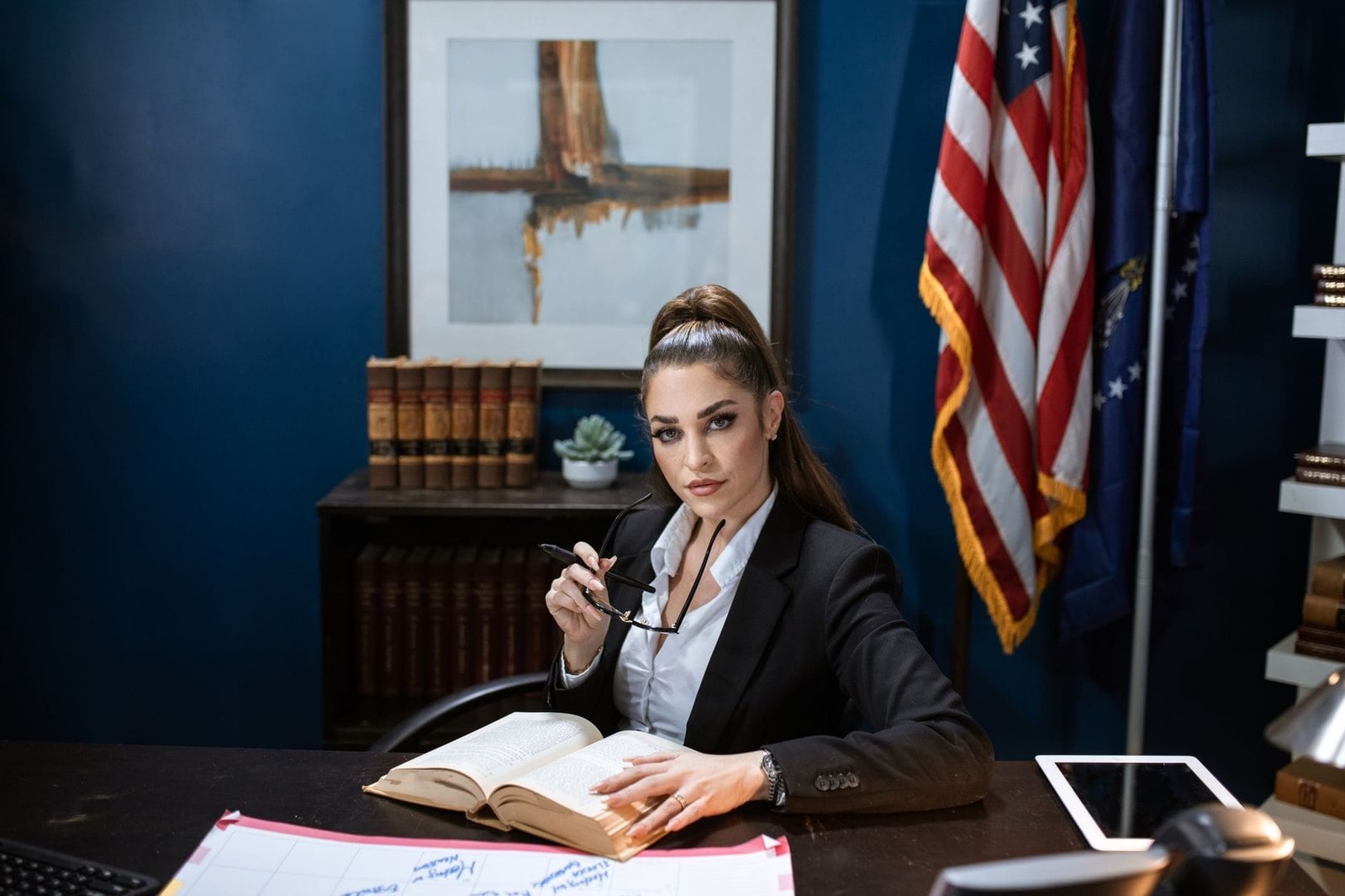 Businesswoman in black blazer and white shirt holds eyeglasses, sitting at a desk with boo