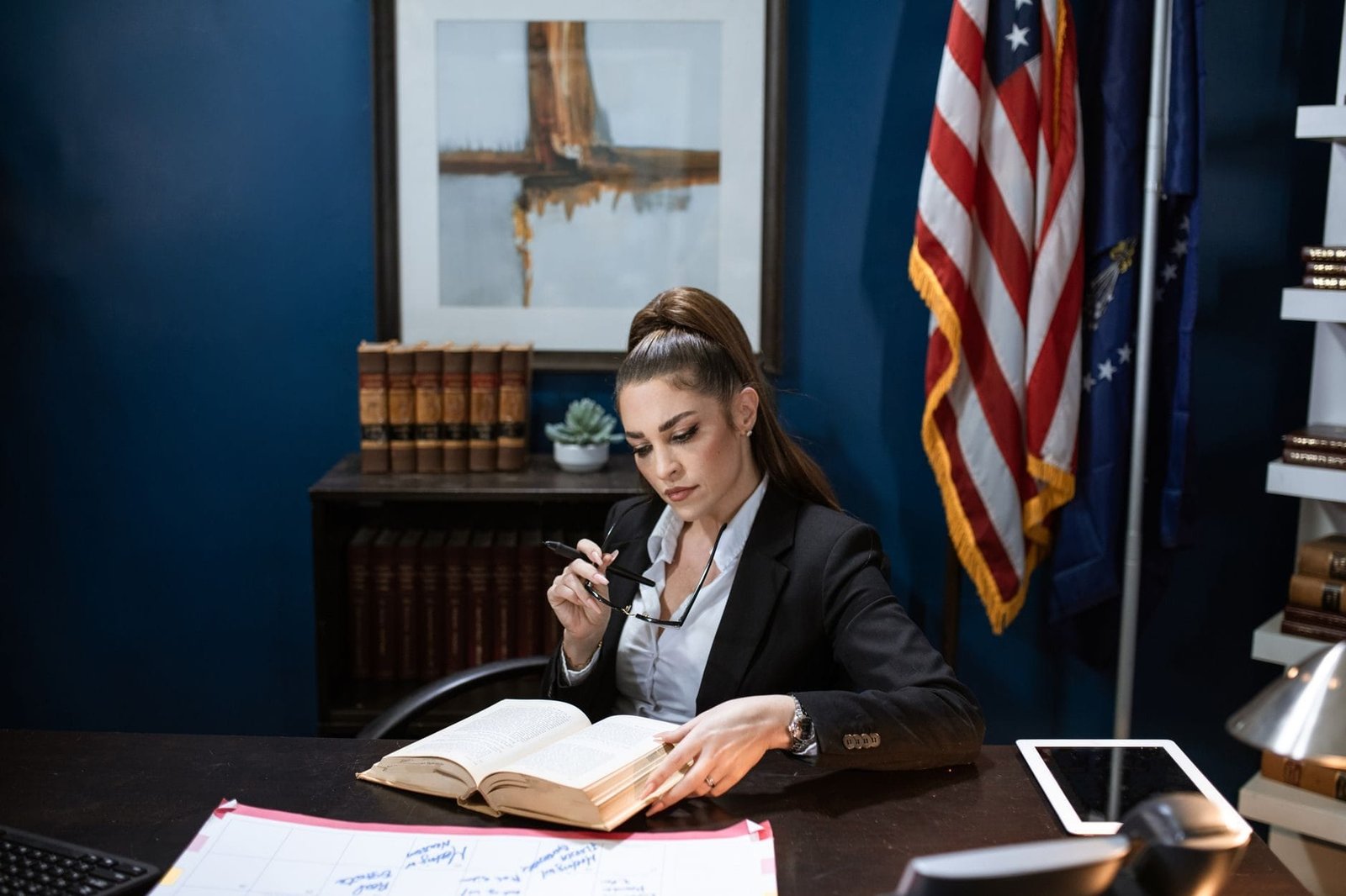 Woman in black blazer reading a book at an office desk, displaying a serious and focused d
