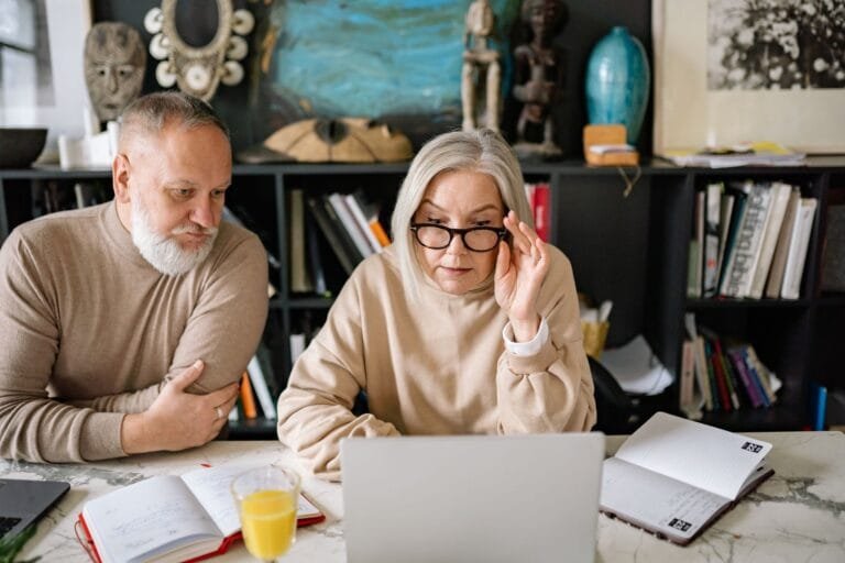 An elderly couple in a cozy home setting engaged with a laptop, planners, and a glass of j