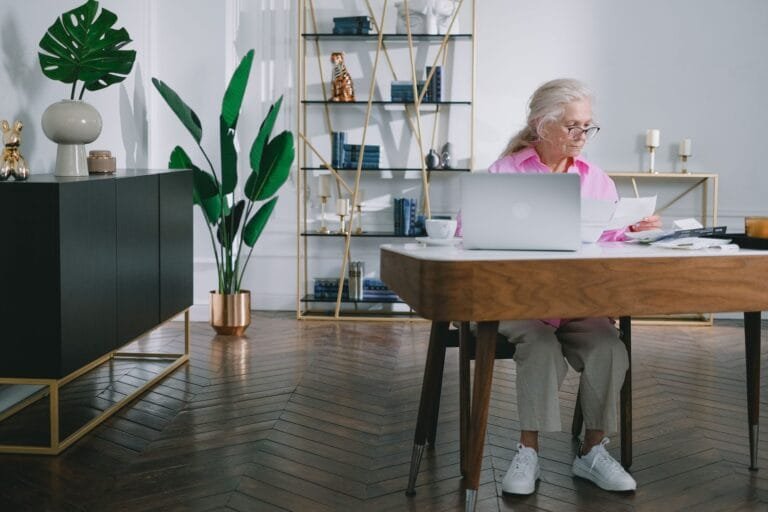 Elderly woman reading papers at a desk with a laptop in a stylish home office setting.