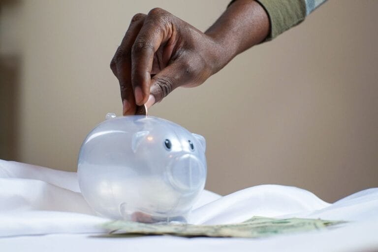 Ressarcimento de descontos do INSS pela MP 1.378/2026: quem recebe e como acompanhar a devolução 5 Close-up of a persons hand placing coins into a transparent piggy bank to save money.