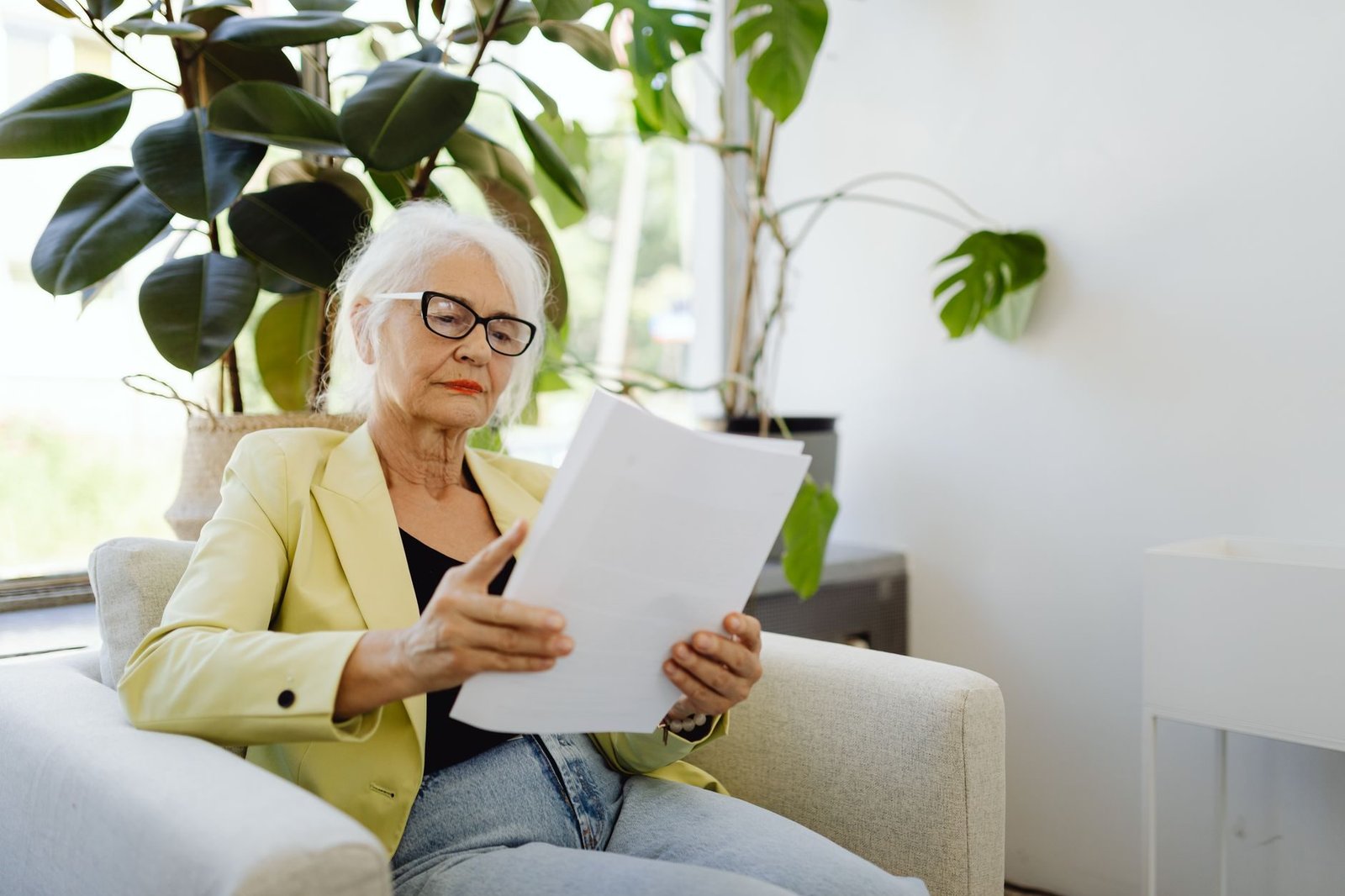 Elderly woman in yellow blazer reading papers at home near plants.