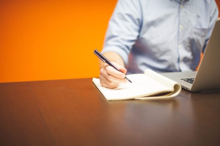 Homem anotando em um caderno ao lado de um notebook, diante de parede alaranjada